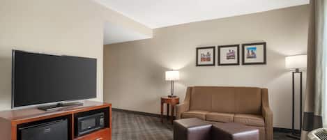 A hotel room with a flat-screen TV, a minibar, a sofa, an ottoman, a lamp, and framed pictures on the wall.
