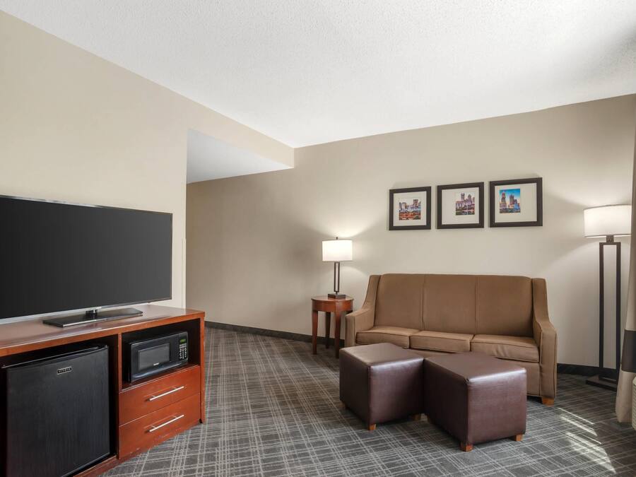 Quarto de hotel com televisão de tela plana, minibar, sofá, banquinho, abajur e quadros na parede.