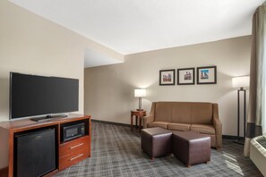 Een hotelkamer met een flatscreen televisie, een minibar, een bank, een pouf, een lamp en ingelijste foto's aan de muur.