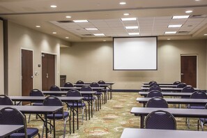 Une salle de conférence avec des rangées de tables et de chaises, un écran de projection et un revêtement de sol en moquette.