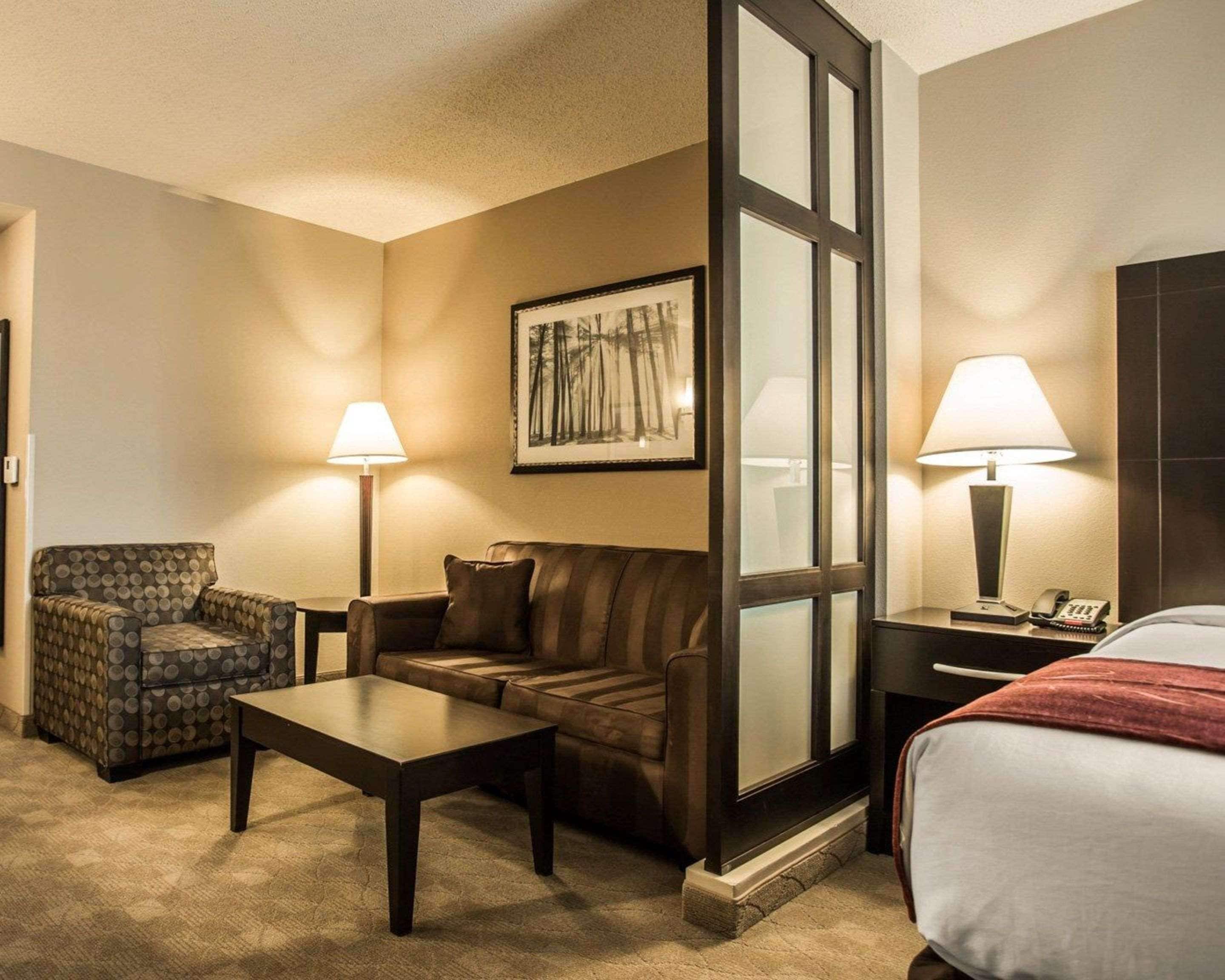 A hotel room with a sofa, armchair, coffee table, and a bed. There are two lamps and a painting on the wall.