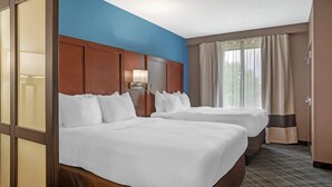 A hotel room with two beds, a wooden headboard, a nightstand with a lamp, and a window with curtains.