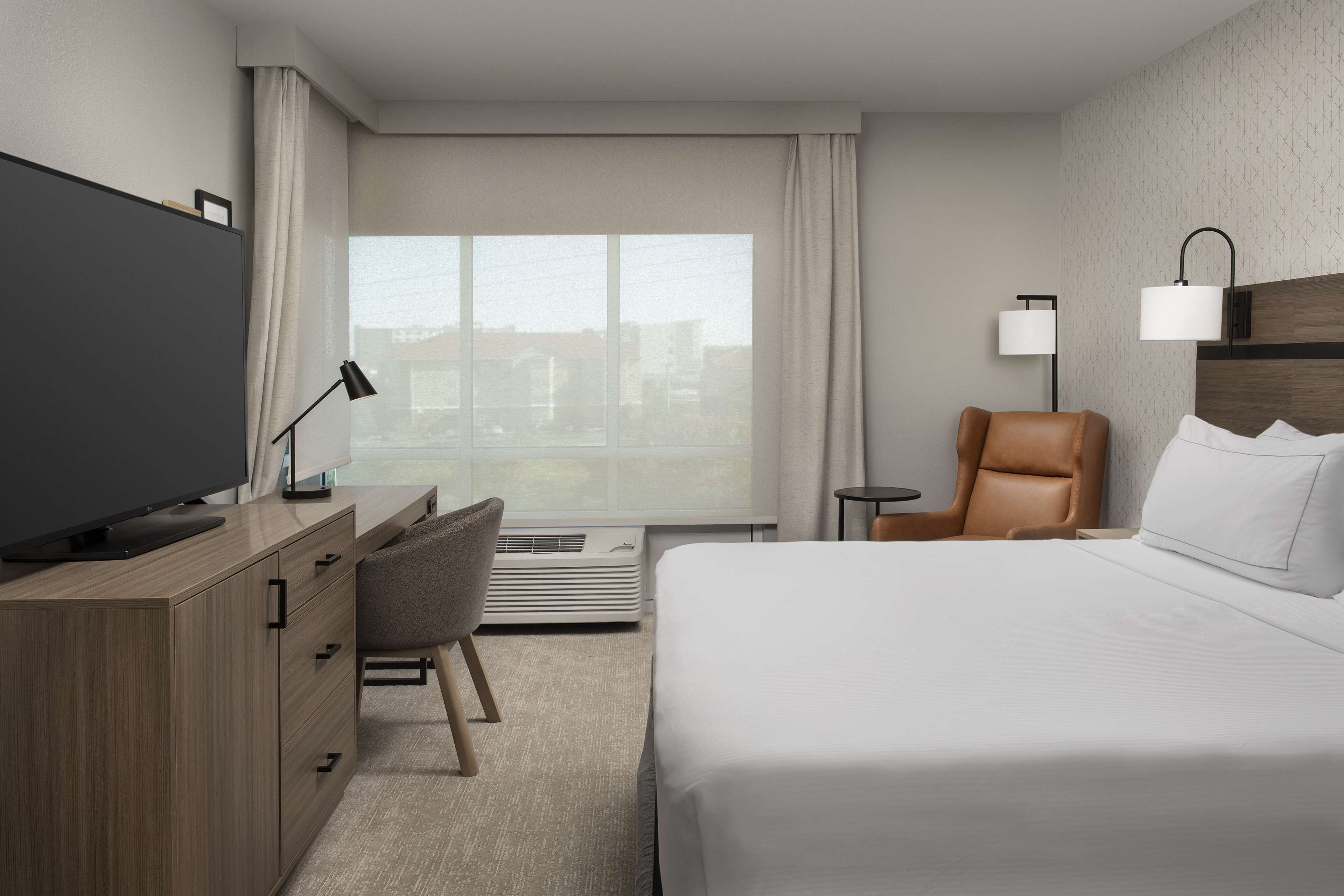 A modern hotel room with a large bed, a desk with a flat-screen TV, a leather chair, and a view of the city through large windows.