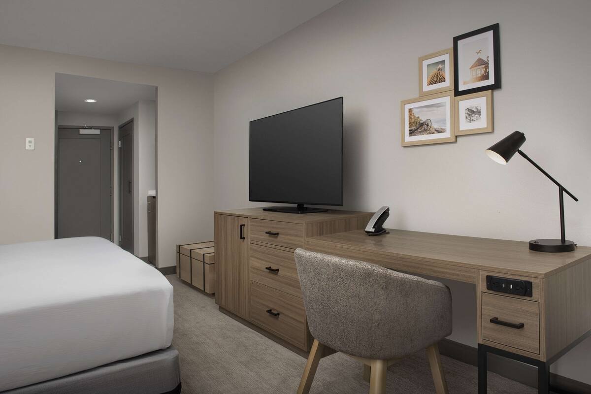 A hotel room with a bed, a wooden desk, a television, a chair, and a desk lamp.