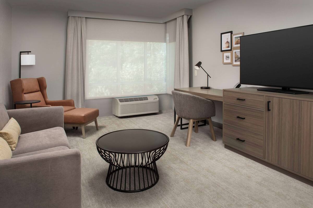 A modern hotel room with a sofa, armchair, round coffee table, desk, television, and a large window with curtains.