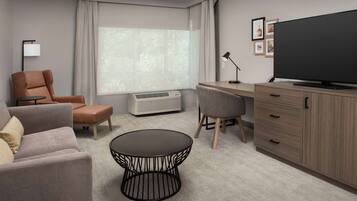Ett modernt hotellrum med en soffa, fåtölj, runt soffbord, skrivbord, tv och ett stort fönster med gardiner.