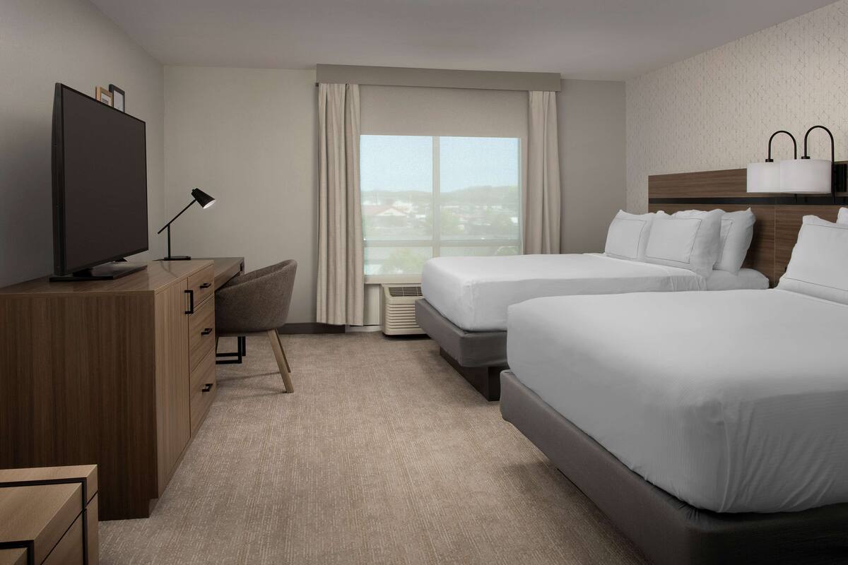 A hotel room with two beds, a desk with a TV, a chair, and a large window with curtains.