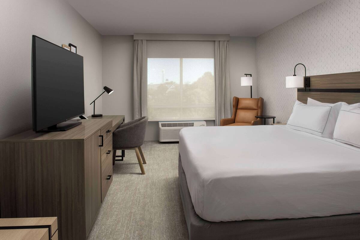 A hotel room with a large bed, a desk with a flat-screen TV, a chair, a lamp, and a window with curtains.