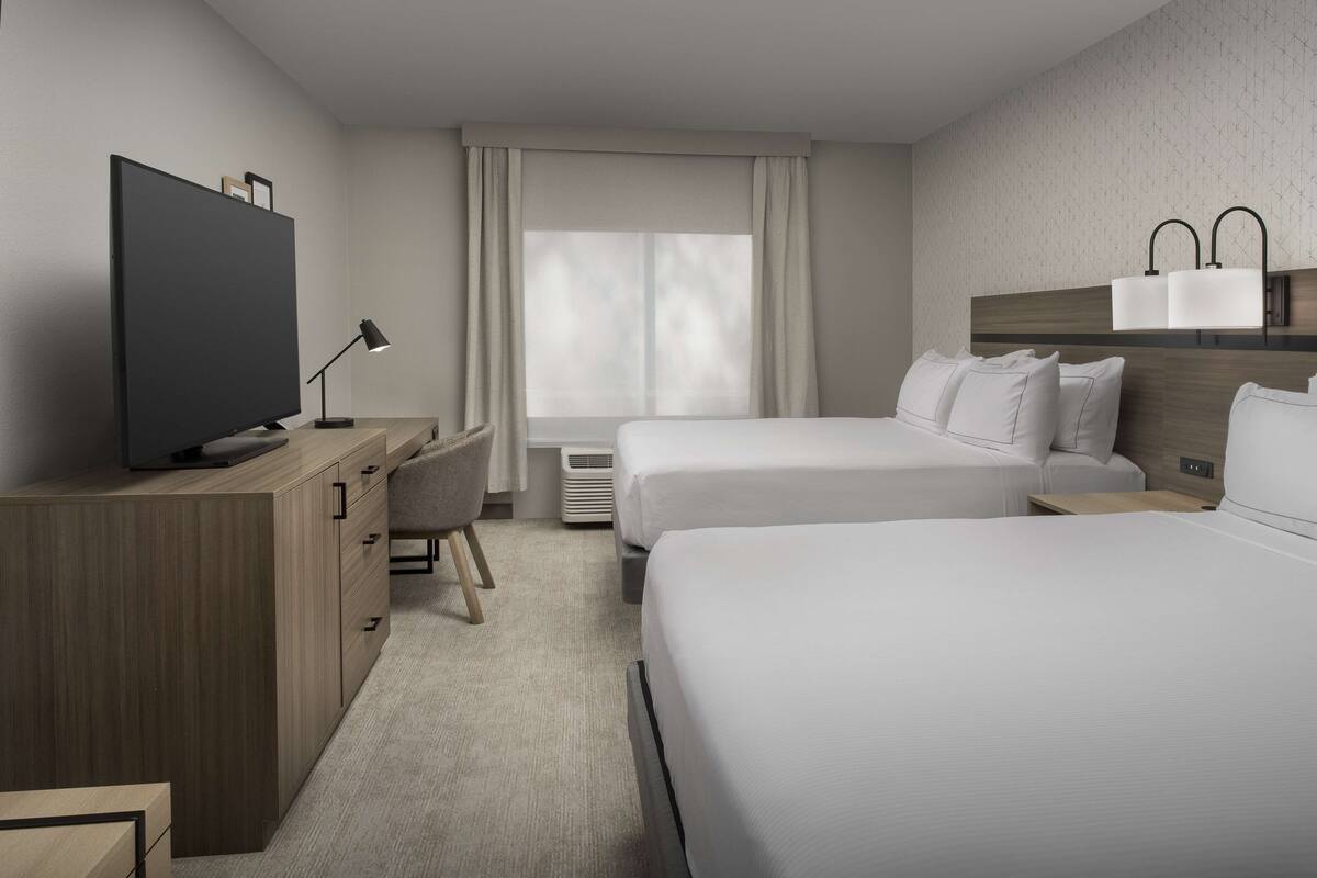 A modern hotel room with two beds, a desk with a flat-screen TV, a chair, and a large window with curtains.