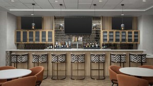 A bar area with a modern design, featuring a large flat-screen TV, wooden cabinets with glass doors, and a variety of liquor bottles. There are bar stools and round tables with orange chairs. - Doubletree by Hilton Chattanooga Hamilton Place (Chattanooga)