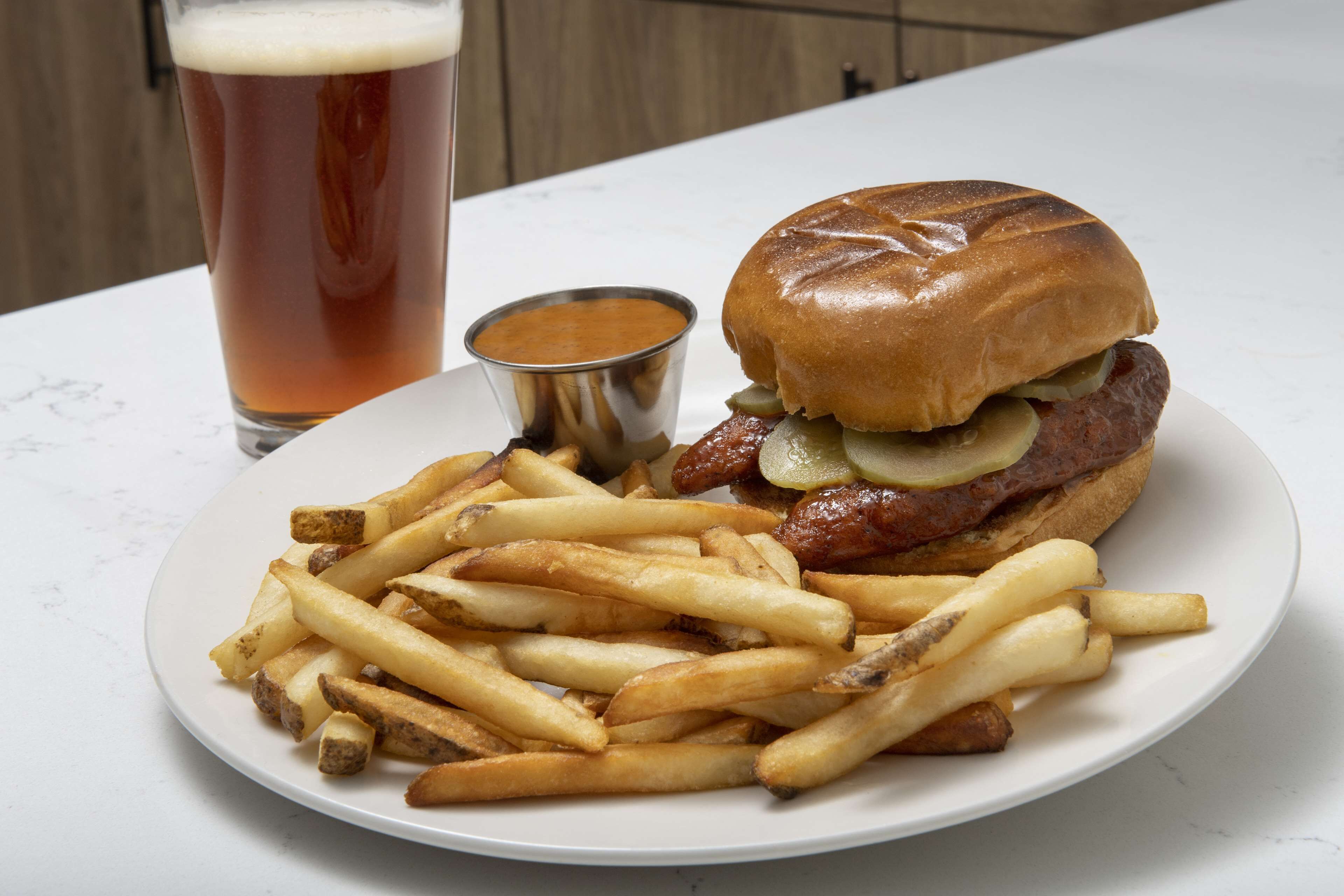 Un plato de papas fritas, una hamburguesa con pepinillos y un vaso de cerveza sobre una mesa blanca.