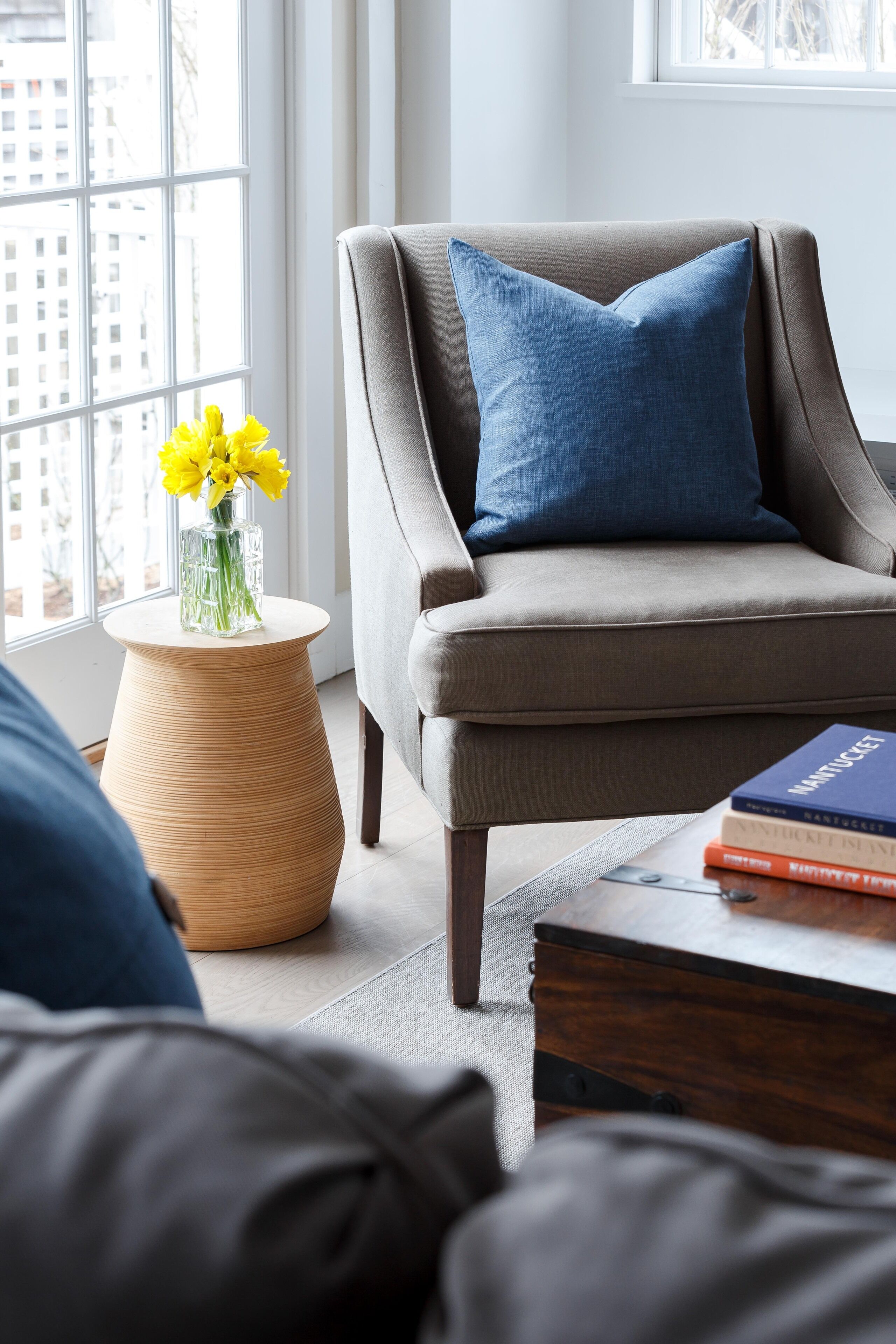 A modern living room with a grey armchair, a round wooden side table with a vase of yellow flowers, and a stack of books on a wooden coffee table.
