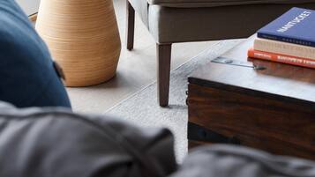 A modern living room with a grey armchair, a round wooden side table with a vase of yellow flowers, and a stack of books on a wooden coffee table.