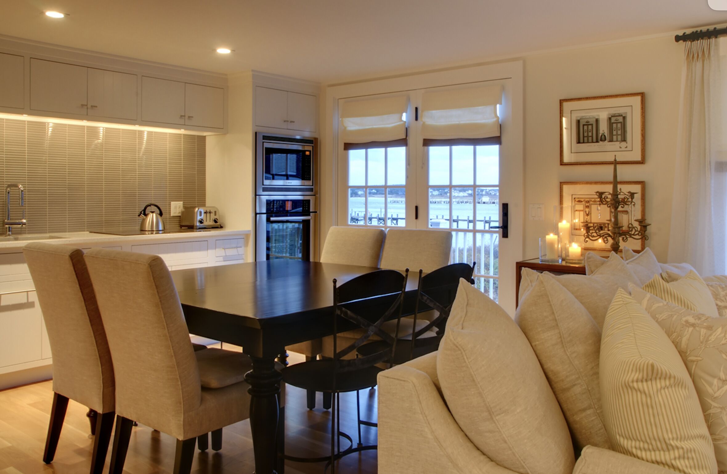 A modern kitchen with white cabinets, a dining table with chairs, and a view through large windows.