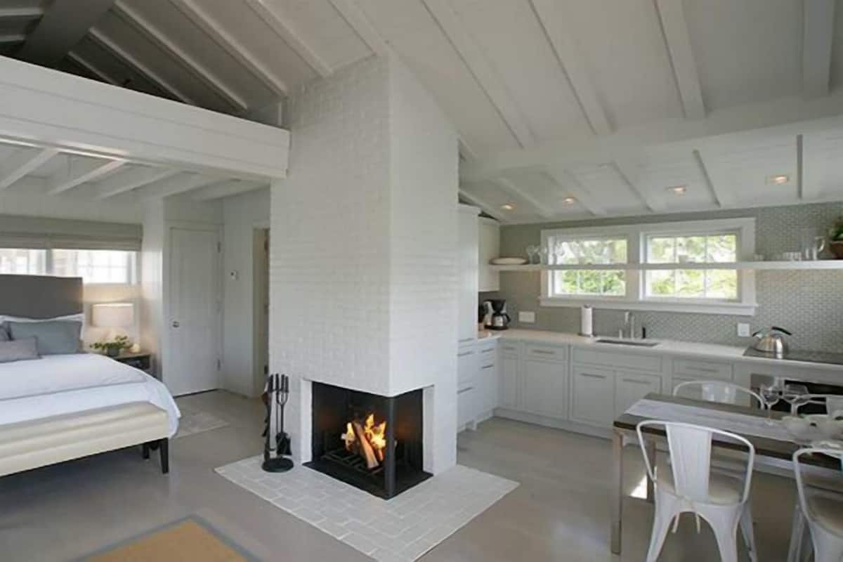 Un dormitorio moderno con una cama grande, una chimenea y una zona de cocina con armarios blancos y una mesa de comedor.