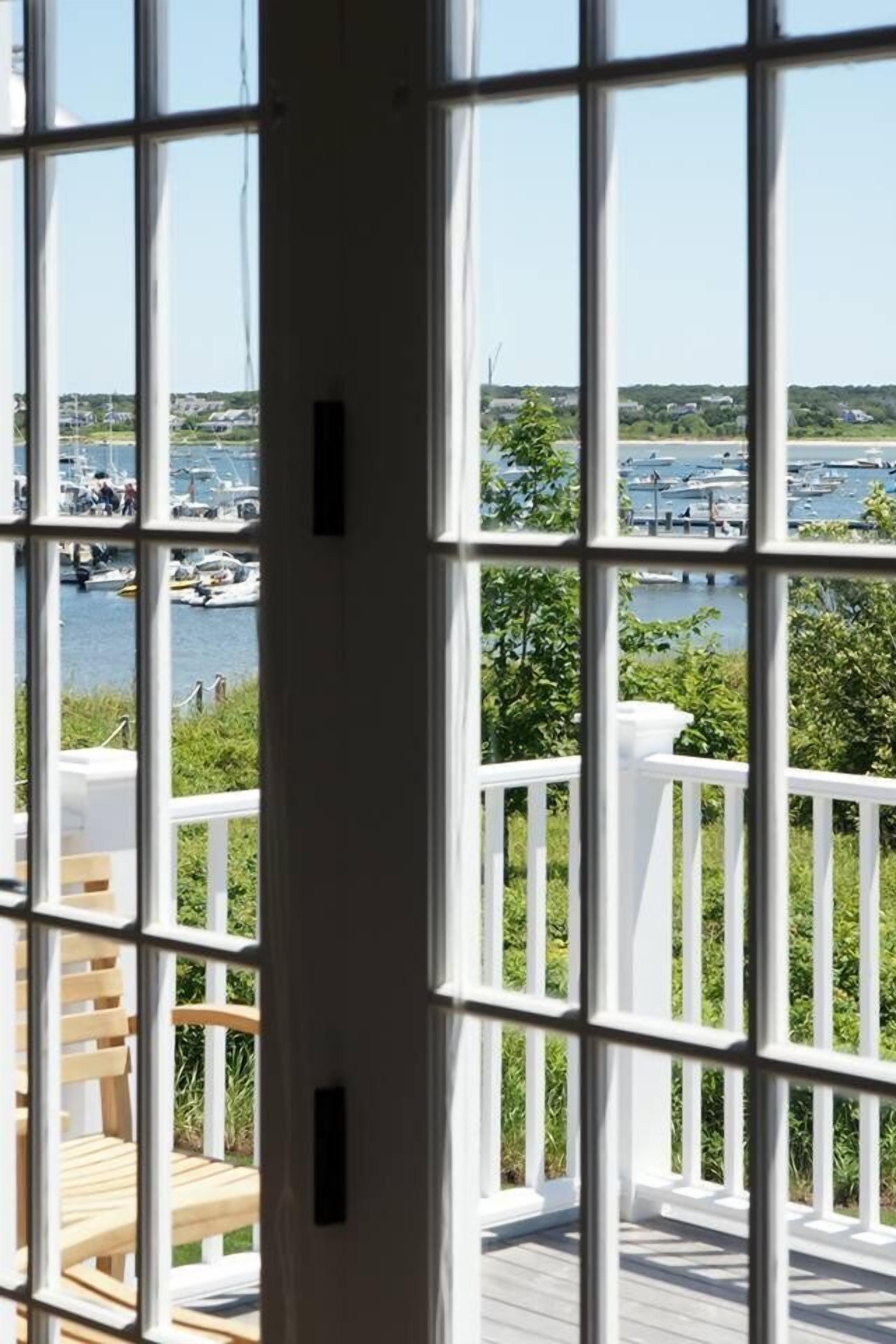 Vista pela janela, com grade branca, sobre um porto com barcos e vegetação.