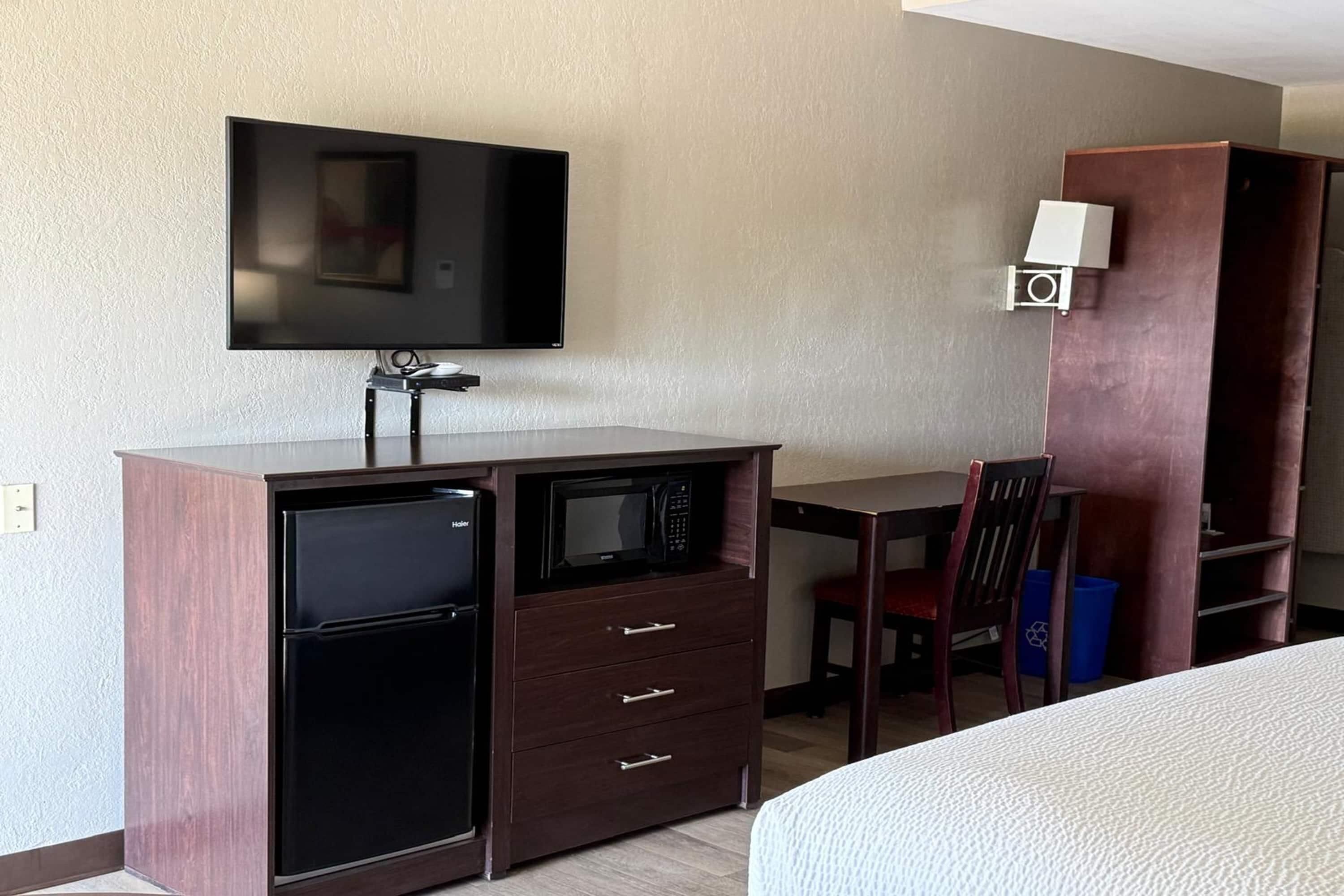 A hotel room with a bed, a desk with a flat-screen TV, a microwave, a refrigerator, a chair, and a bookshelf.