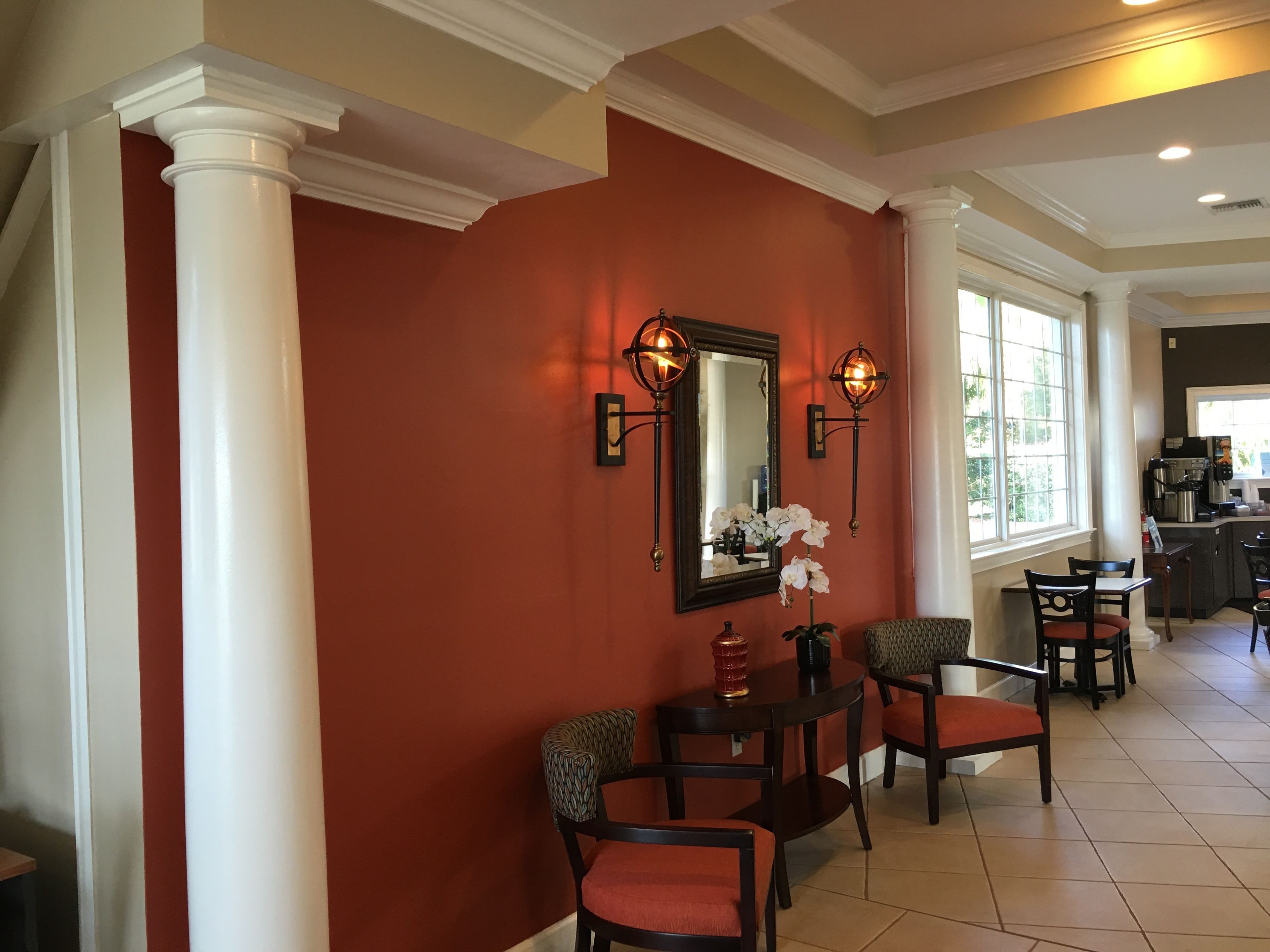 A hotel lobby with a red wall, white columns, a mirror, two wall sconces, a vase with flowers, and a seating area with chairs and a table.