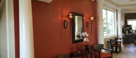 A hotel lobby with a red wall, white columns, a mirror, two wall sconces, a vase with flowers, and a seating area with chairs and a table.