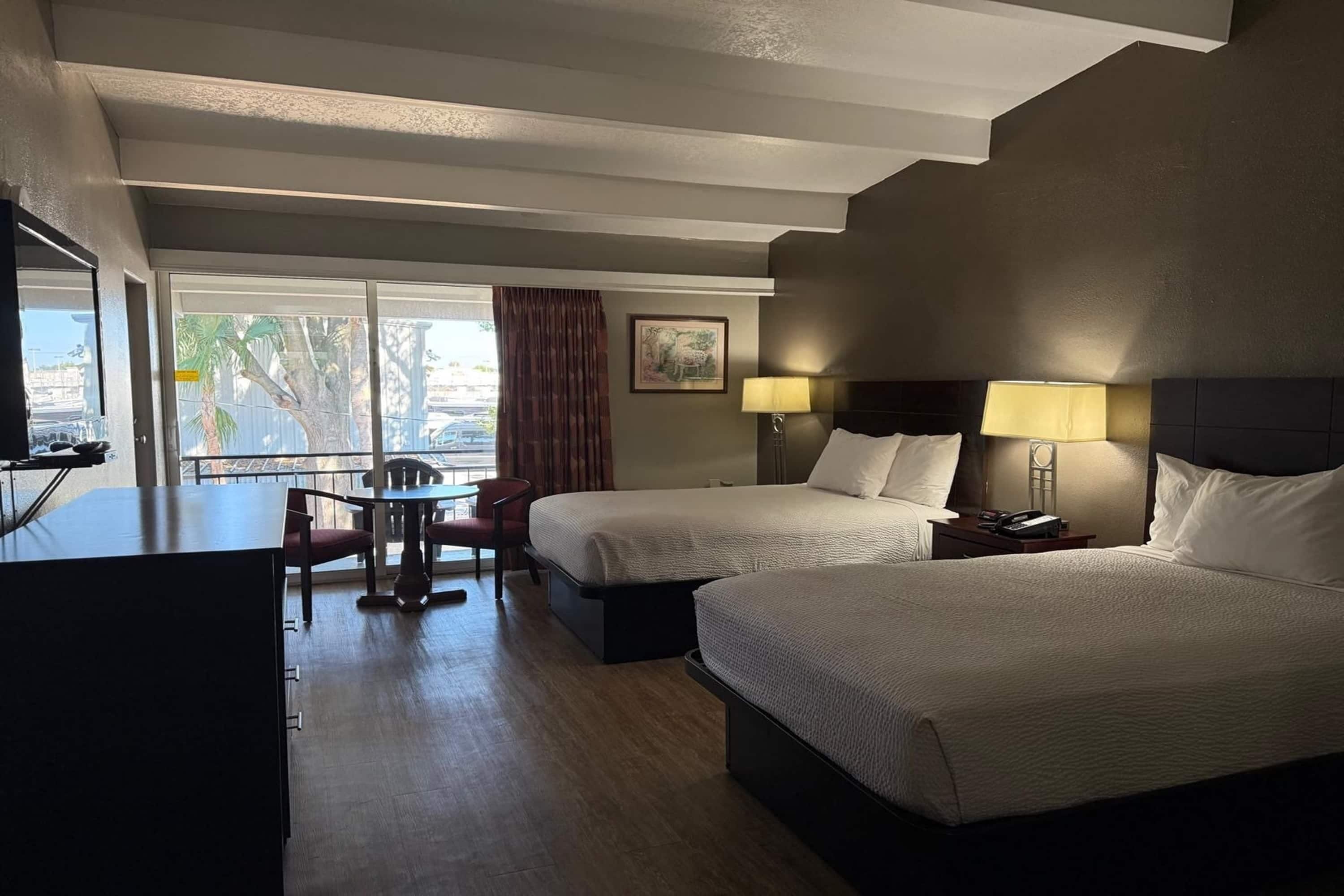A hotel room with two beds, a desk, a chair, a table, and a view of palm trees.