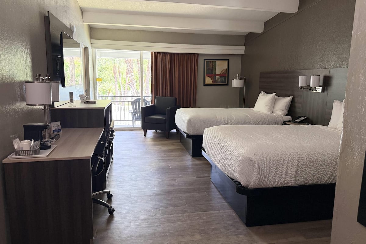 Habitación de hotel con dos camas, un escritorio con computadora, una silla, un televisor y una ventana con cortinas.