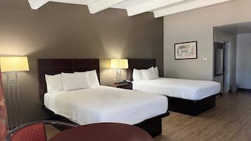 A hotel room with two beds, a bedside table with a lamp, a chair, and a small table.