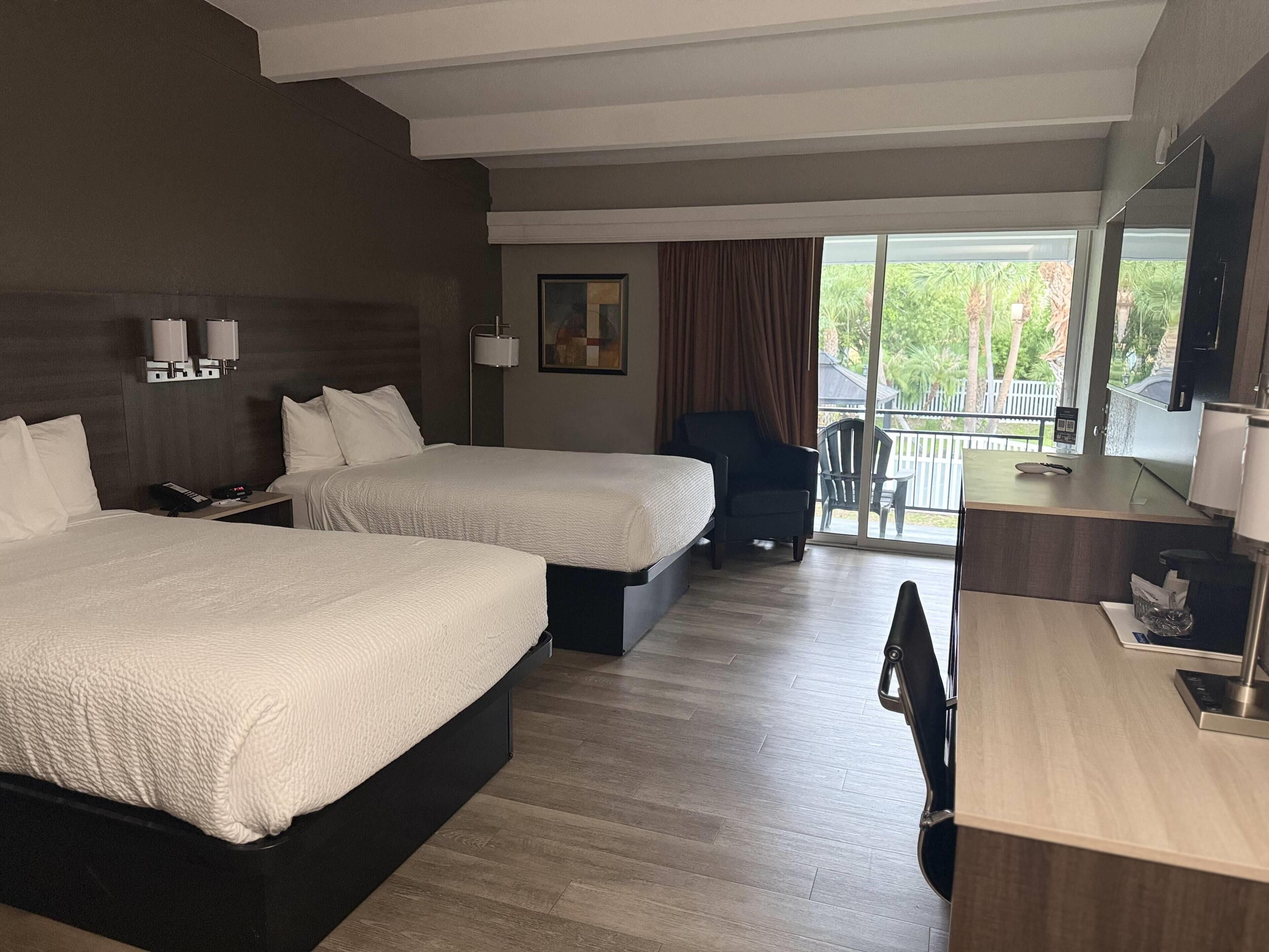 A hotel room with two beds, a desk, a chair, a balcony with a view, and a painting on the wall.