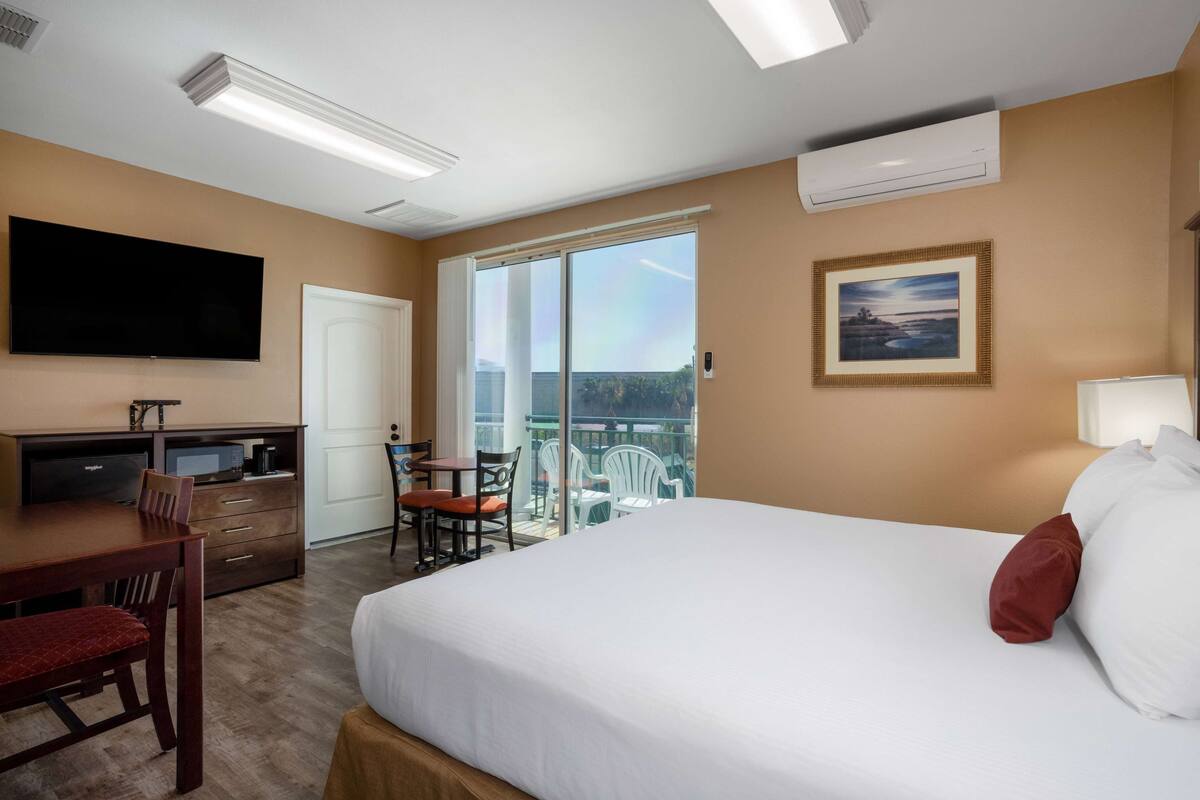 Habitación de hotel con cama, escritorio con televisor, balcón con sillas y un cuadro en la pared.