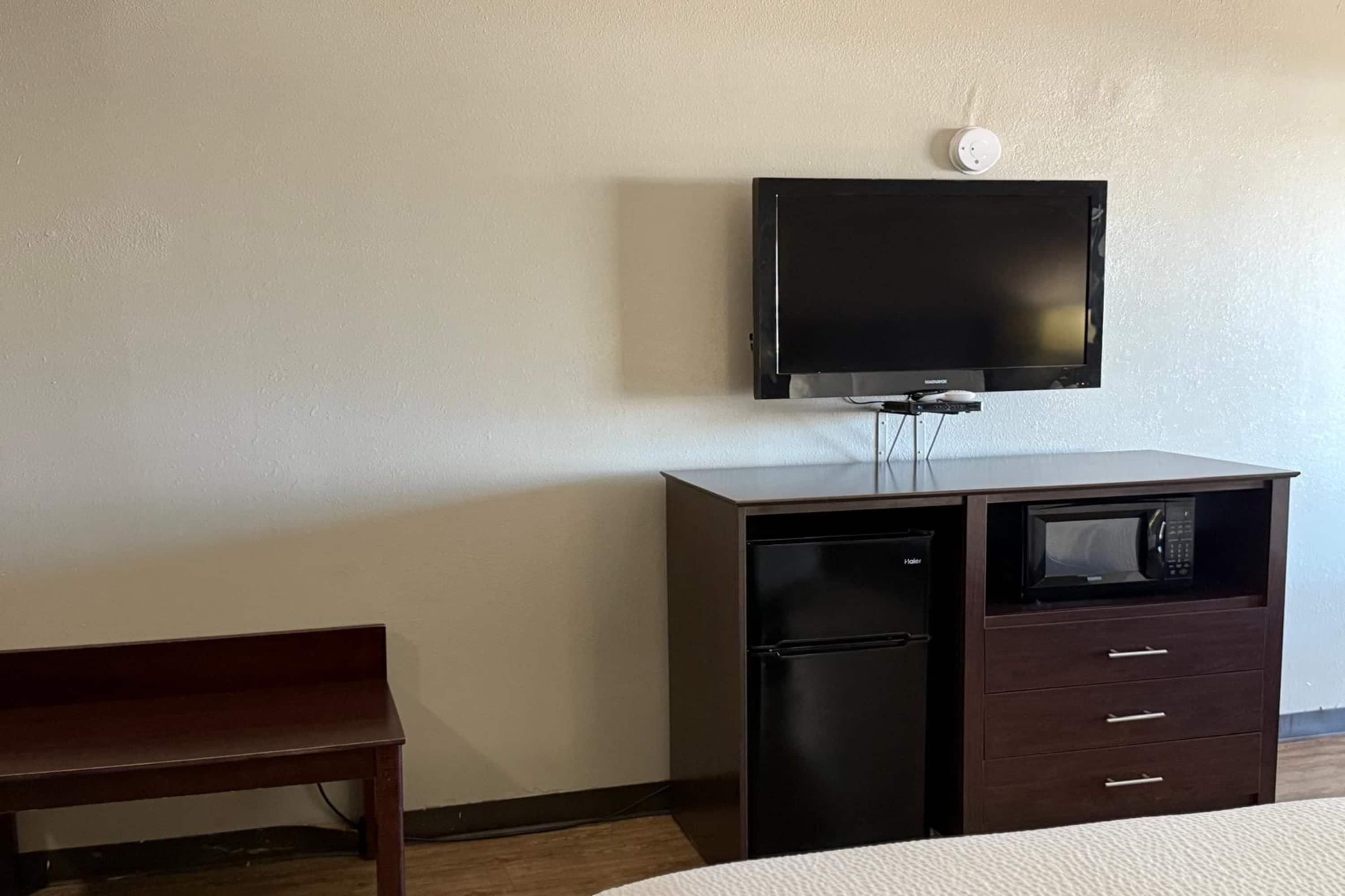 A room with a bed, a desk with a television, a microwave, and a bench.