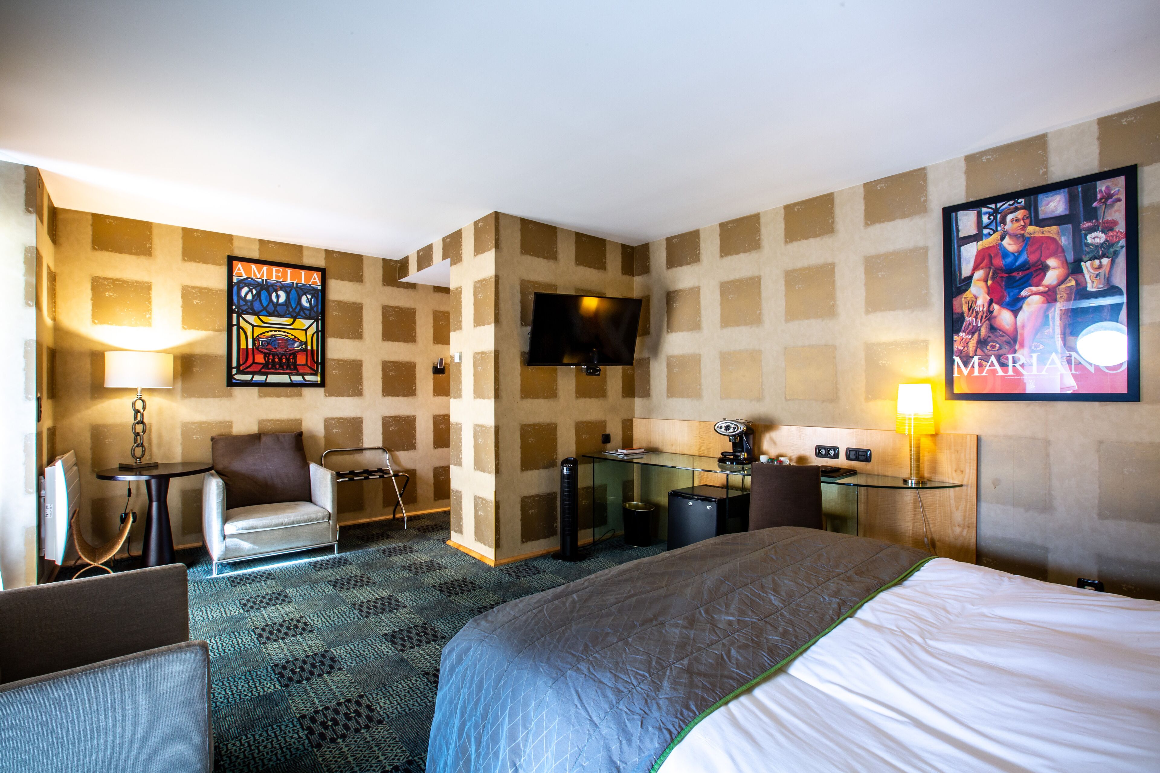 A hotel room with a bed, a desk, a chair, a TV, and a painting on the wall.