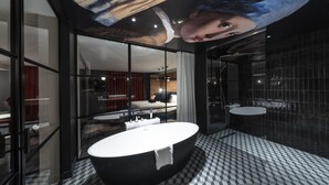 Een moderne badkamer met een groot bad, een douchecabine van glas en uitzicht op een slaapkamer met een bed en nachtkastje.