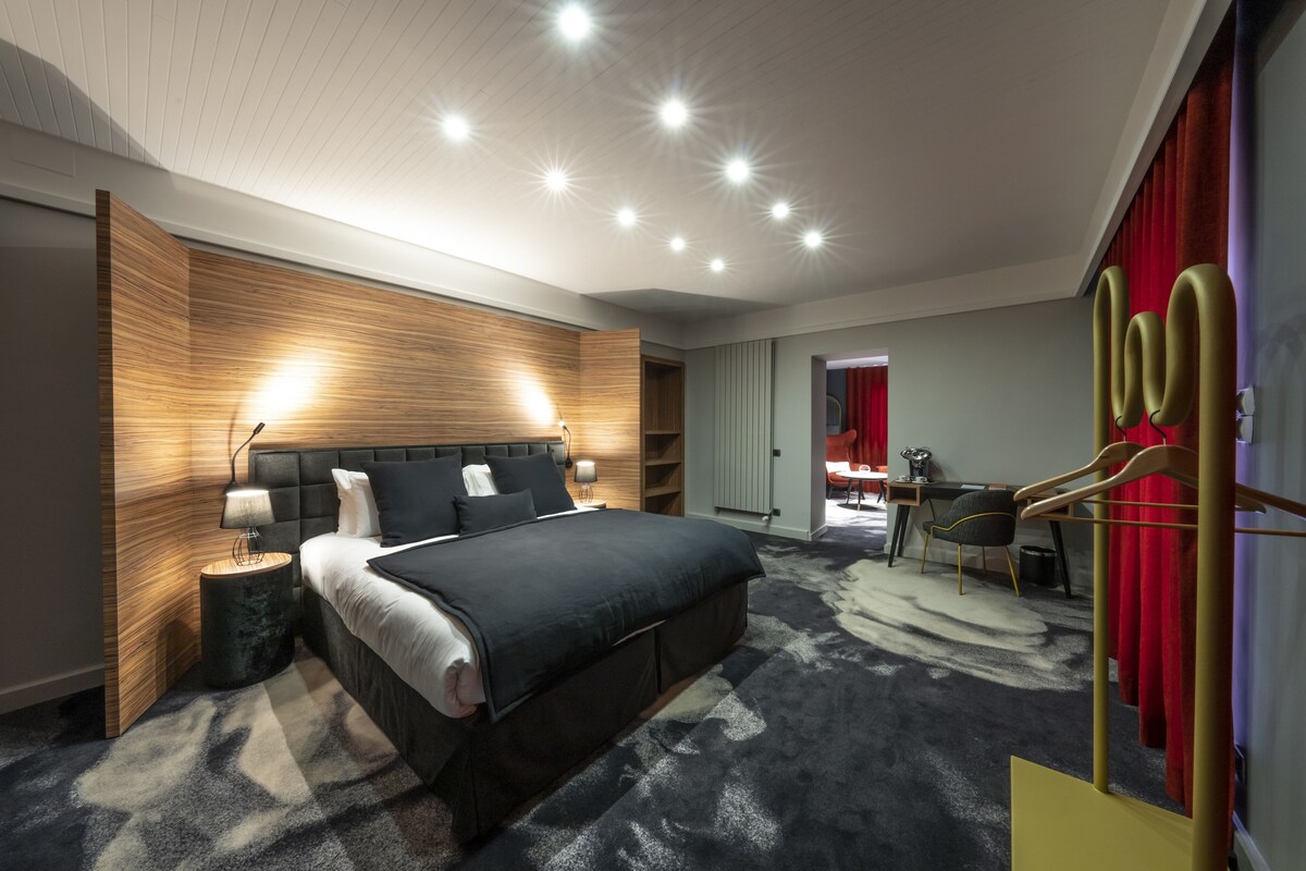 A modern hotel room with a large bed, bedside lamps, a wooden accent wall, a desk with a chair, and a red curtain.