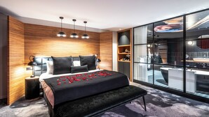 Een moderne slaapkamer met een groot bed, nachtkastjes, een bankje en een glazen wand waardoor een badkamer en keuken zichtbaar zijn.