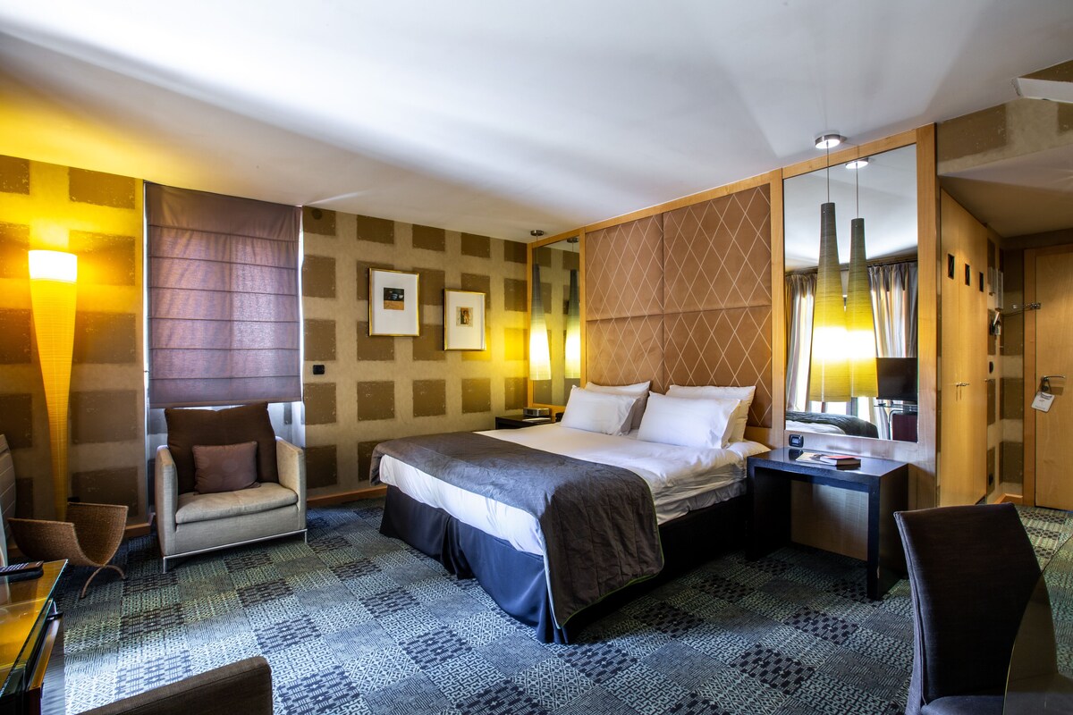 A hotel room with a large bed, a sitting area with a chair, a desk, and a mirror.
