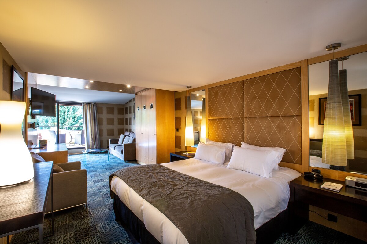 A modern hotel room with a large bed, a desk, a chair, and a view of the outdoors.