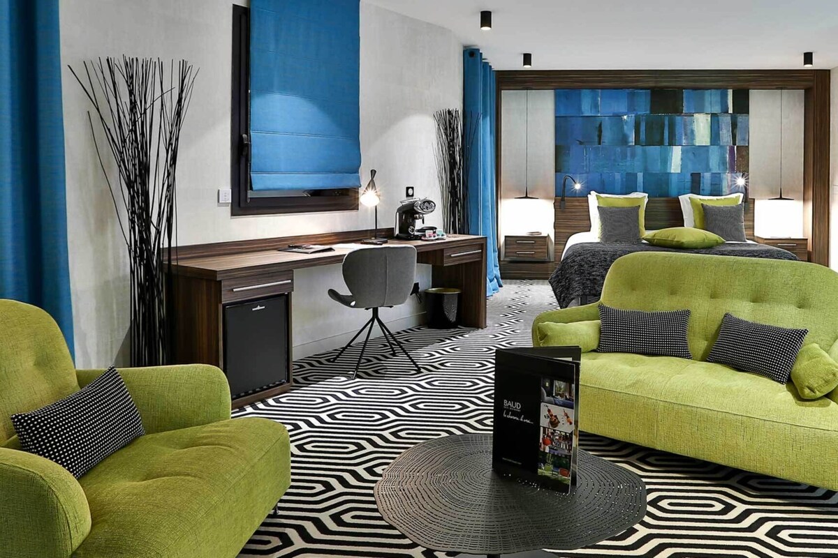 A modern hotel room with a geometric patterned floor, a desk with a computer, two green armchairs, a large mirror, and a view of the city through a window.
