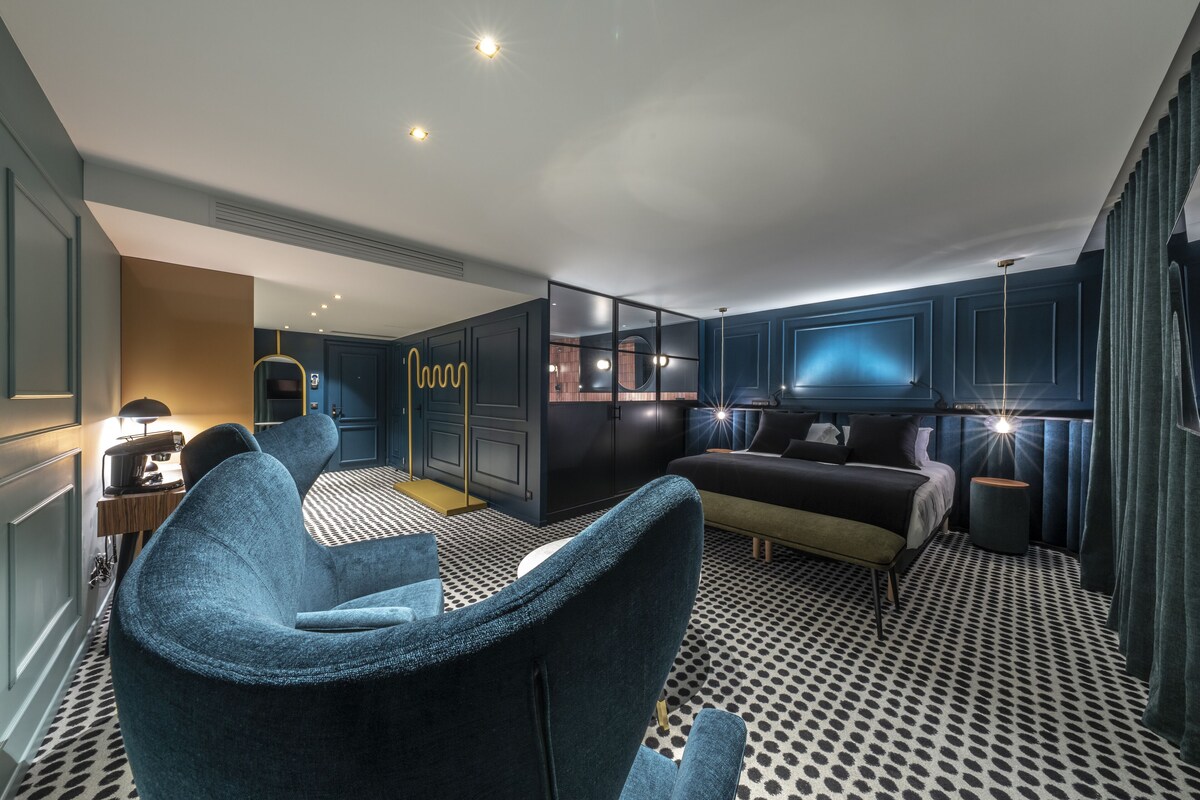 A modern hotel room with a sofa, a bed, a TV, and a desk with a lamp.