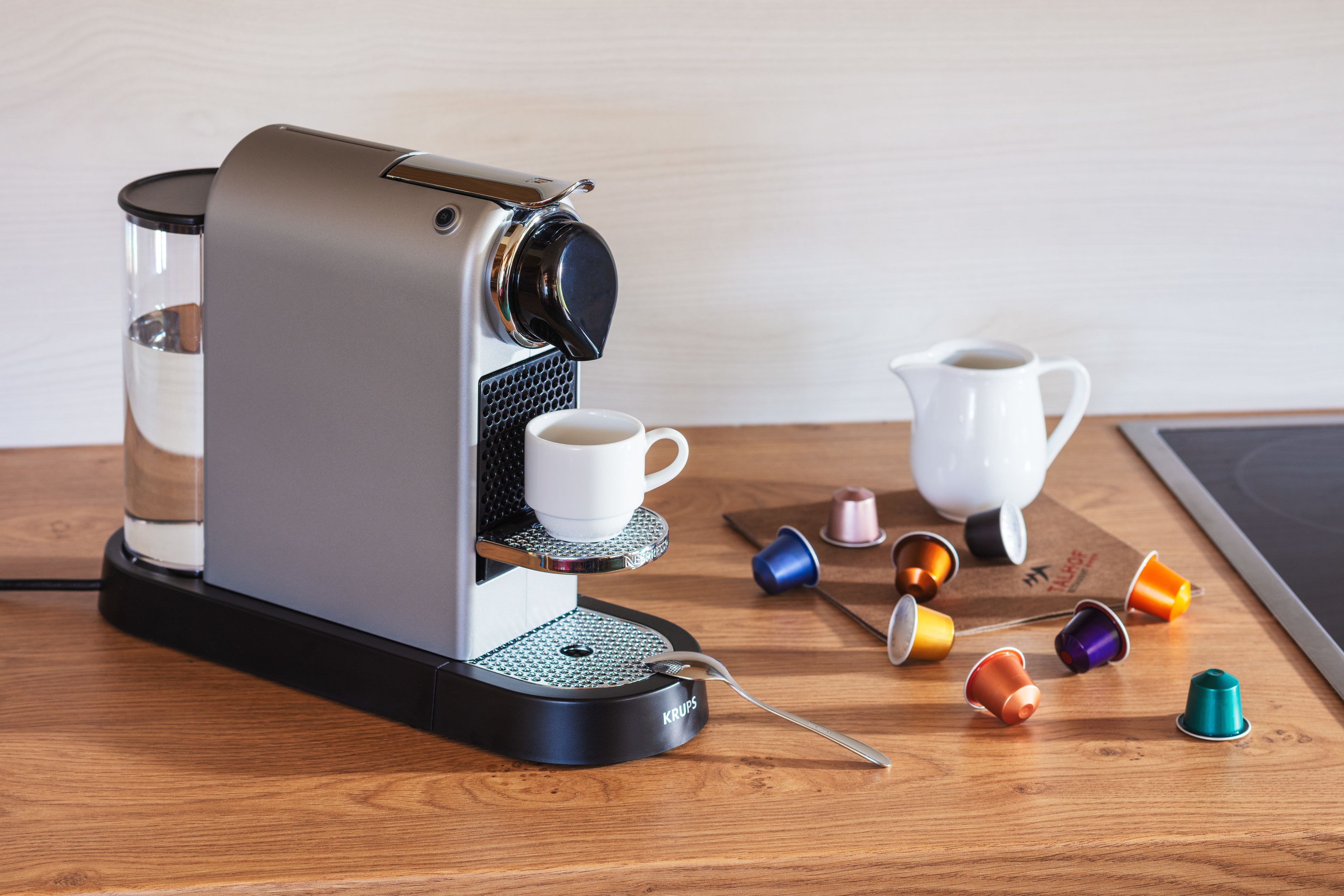 Una cafetera con portacopas, una jarra y cápsulas de café sobre una mesa de madera.