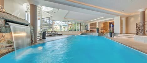 An indoor swimming pool with a waterfall feature, a staircase, and lounge chairs.