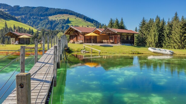 A wooden bridge leading to a cabin with a view of a clear lake and mountains. - Hotel Talhof (Wildschonau)
