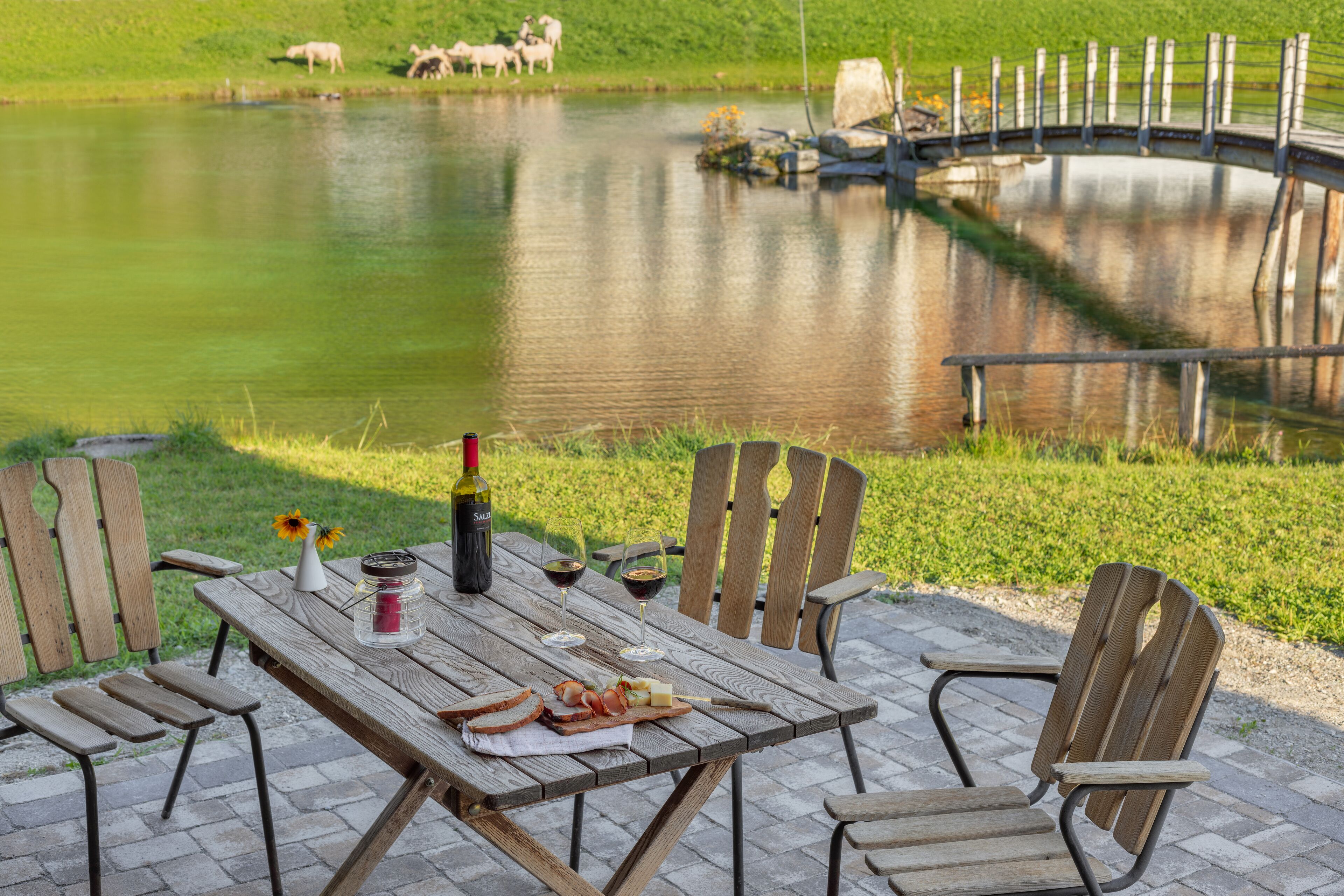 Ein Holztisch draußen mit Stühlen, einer Weinflasche, Gläsern und einer Käseplatte.