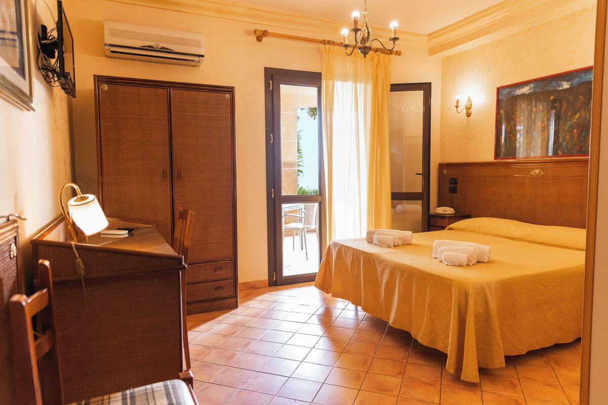 A hotel room with a bed, a desk, a chair, a wardrobe, and a door leading to an outdoor area.