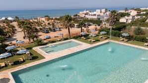 A large rectangular pool with clear blue water, surrounded by lounge chairs and umbrellas, with a view of the sea and buildings in the background. - Triscinamare Hotel Residence (Castelvetrano)