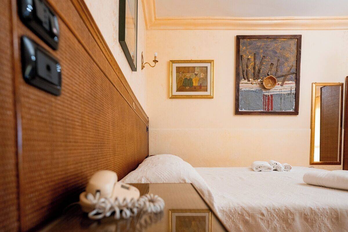 A hotel room with a wooden headboard, a bed with white linens, two framed artworks on the wall, and a bedside table with a telephone and a folded towel.