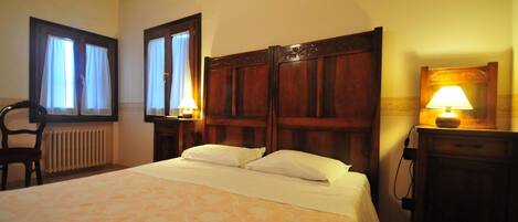 Habitación de hotel con una cama grande, cabecera de madera, dos lámparas de noche, una silla y una ventana con cortinas.
