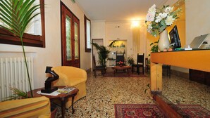 Un salón acogedor con un sillón amarillo, una mesa de centro de madera y un tapete rojo con estampado. Hay una planta en maceta, un jarrón con flores y un ordenador portátil sobre un escritorio de madera.