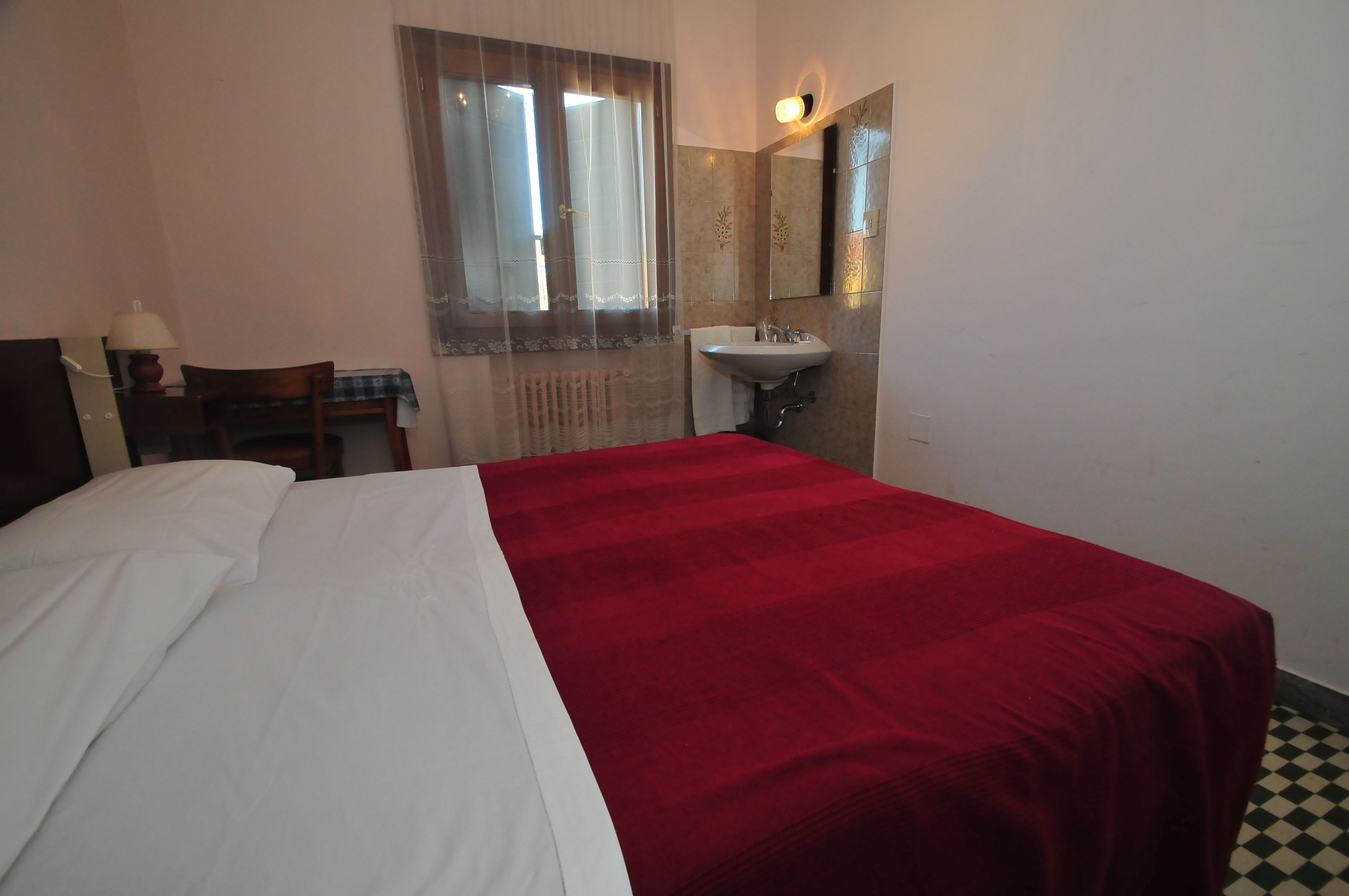 A hotel room with a bed, a desk with a lamp, a window with curtains, a sink, and a mirror.