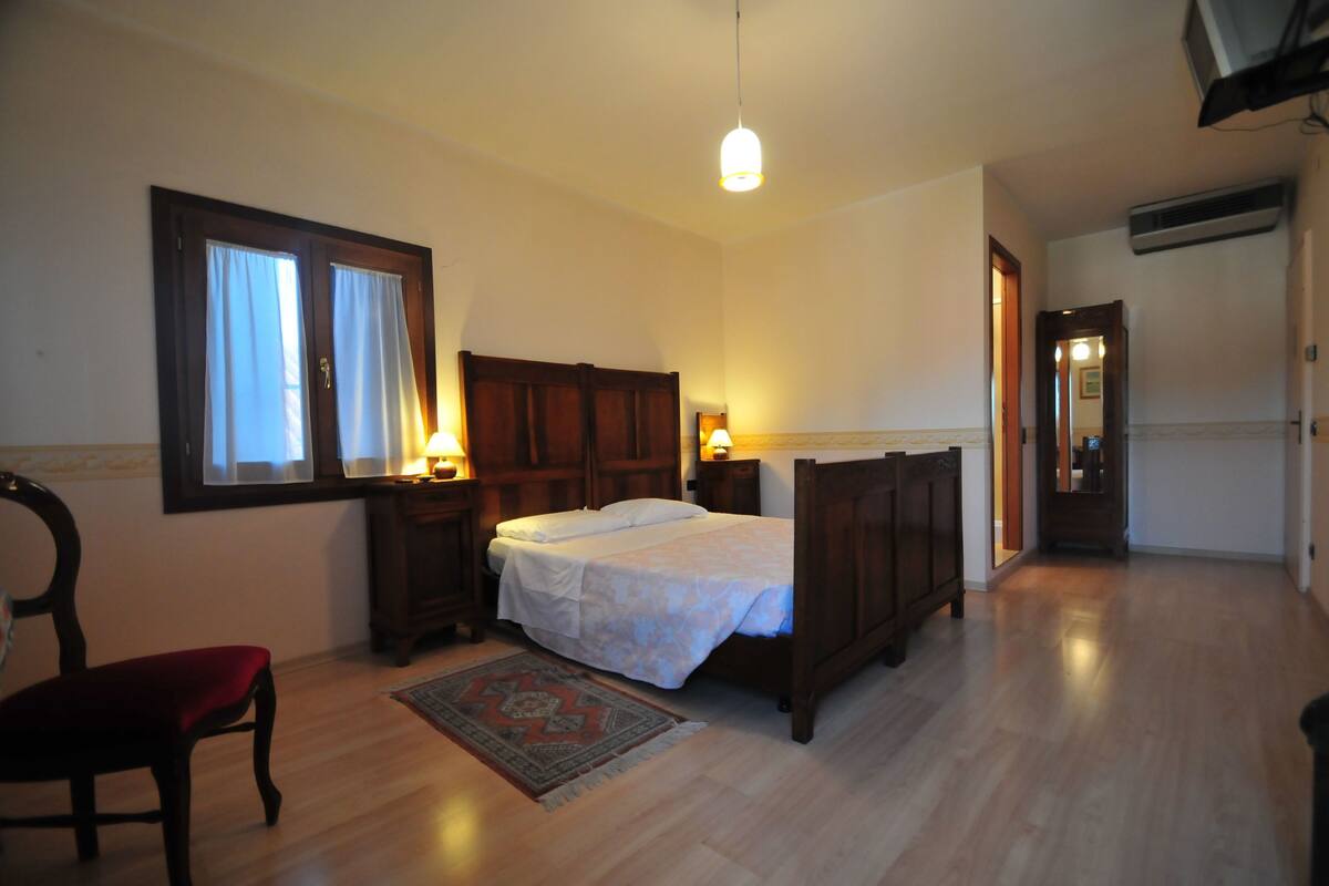 A bedroom with a wooden bed, a chair, a nightstand, a window with curtains, and a hanging light fixture.
