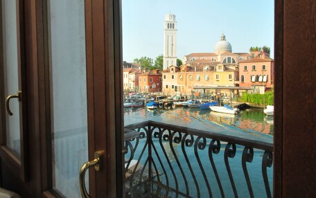 A view from a window with a balcony, overlooking a canal with boats, historic buildings, and a tower.