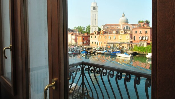Vista desde una ventana con balcón, sobre un canal con barcos, edificios históricos y una torre.